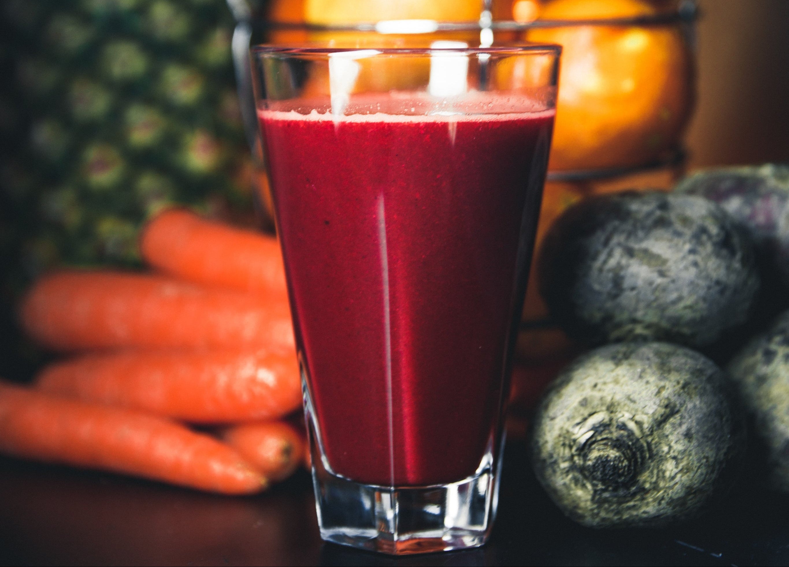 Glass of red juice with oranges, carrots, and beets on a dark background